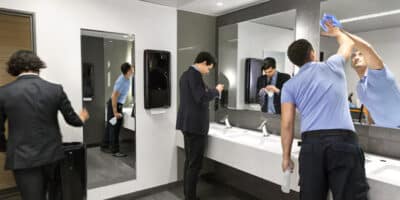 3 men in a public restroom where Tork Peakserve towel dispensers are being used. One is a maintenance man cleaning the mirror. Another is cleaning a stain off his necktie. A third is discarding a paper towel as he exits the bathroom.
