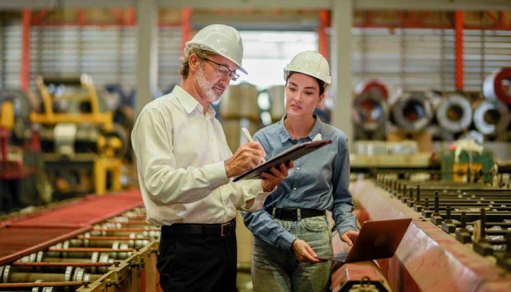 Woman and man in a factory setting up predictive maintenance processes on a tablet and a laptop. 